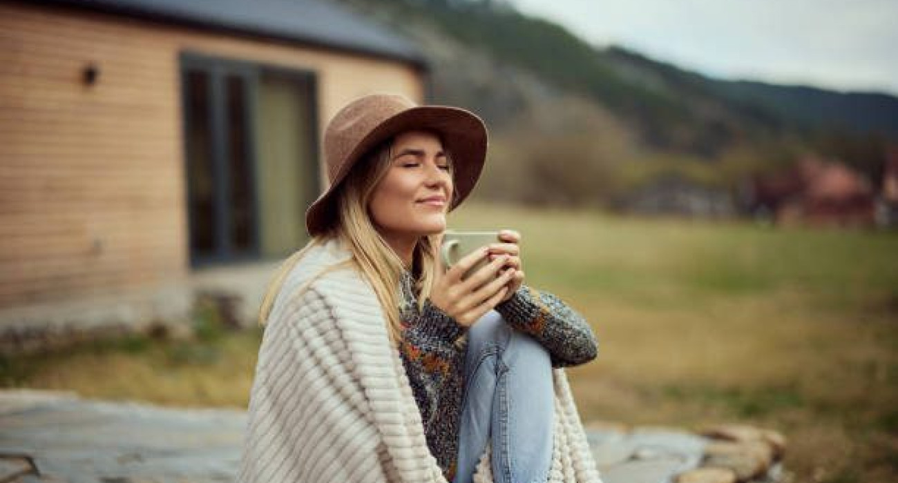 A smiling girl holding a cup of coffee, sitting outdoors, wearing a hat, wrapped in a blanket.